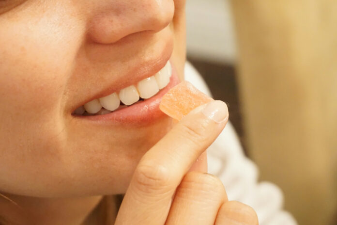 cannabis gummies grow up Close-up profile of a smiling woman holding a single square peach-colored cannabis gummy candy to her lips.