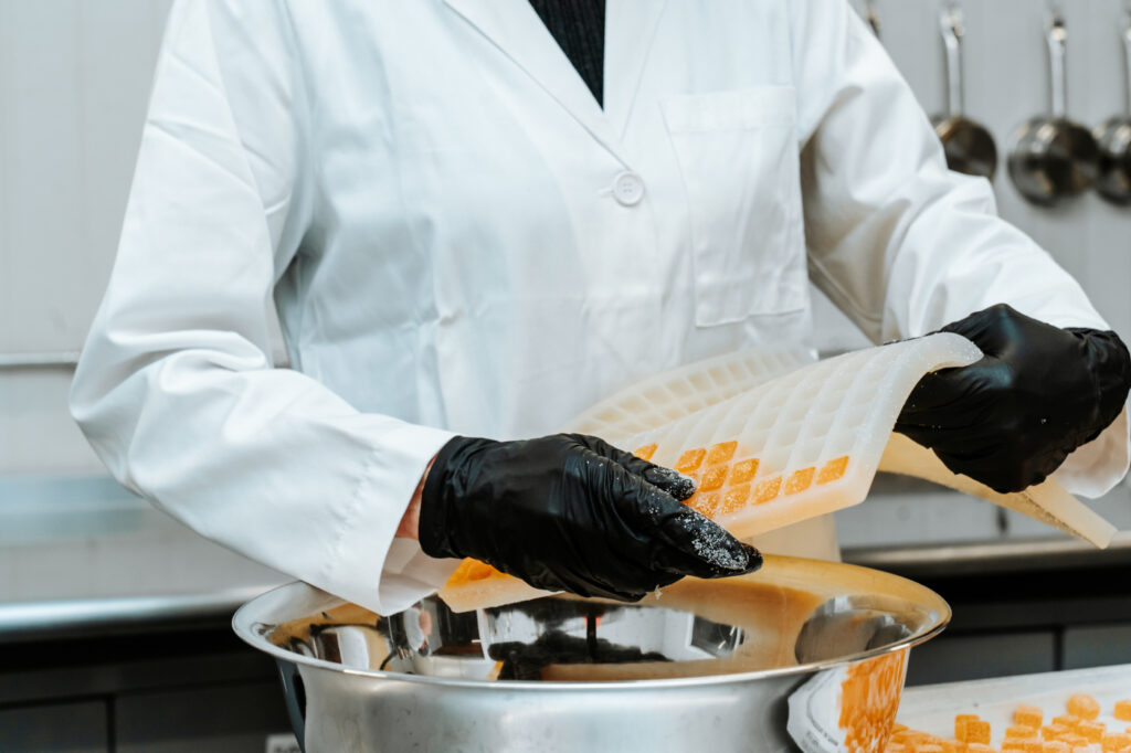 A person in a white lab coat and black gloves peeling a flexible silicone mold to release orange gummy candies into a large stainless steel bowl.