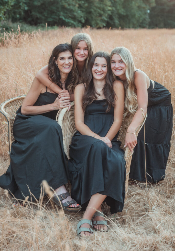 Four women dressed in long black dresses sit closely together in a field of tall, dry grass. The natural setting and warm light create a contemplative, intimate mood that evokes reflection, loss, and generational strength.