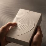 Hands hold an unbranded textured package as ripples radiate from a fingertip.
