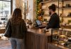Retail employee uses a touchscreen POS while serving a customer at a wood checkout counter.